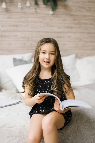 Κομψό κορίτσι πέντε ή έξι ετών με μαύρο φόρεμα με πούλιες να στριφογυρίζουν και να διαβάζουν ένα βιβλίο ή περιοδικό - Φωτογραφία, εικόνα