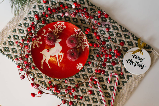 Karácsonyi üdvözlőlap, szilveszteri lakoma. Piros kerámia lemez cukorral vörös alma, karácsonyfa játék, cukornád és ágak piros bogyók - Fotó, kép