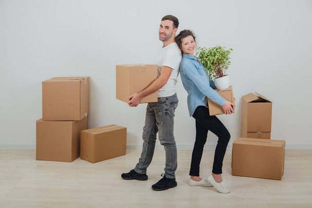 Happy wife and husband holding carton boxes, standing back to back, smiling extremely satisfied to move into new apartment. - Фото, изображение