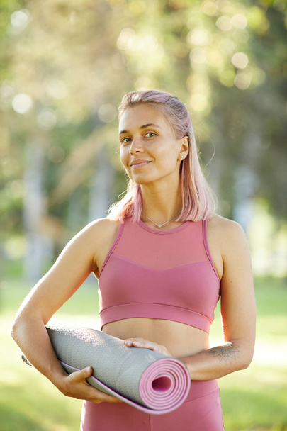 Portrait of beautiful healthy woman in pink top holding exercise mat and smiling at camera while standing outdoors in the park - Fotoğraf, Görsel