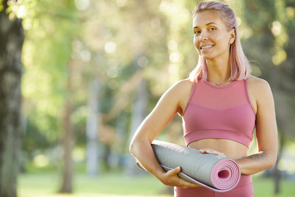 Портрет счастливой молодой женщины в розовом топ проведение упражнения мат и улыбаясь она собирается тренироваться в парке
 - Фото, изображение