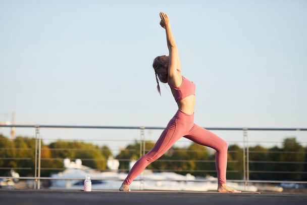 Jeune femme en vêtements de sport rose debout et les mains tendues vers le haut pratiquant le yoga dans la ville
 - Photo, image