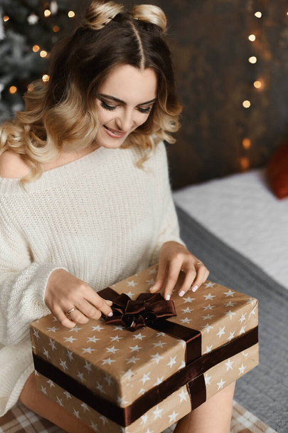 Piękny model dziewczyna z modną fryzurą w oversize sweter siedzi w pobliżu choinki z prezentem świątecznym i pozowanie we wnętrzu urządzone na Nowy Rok - Zdjęcie, obraz