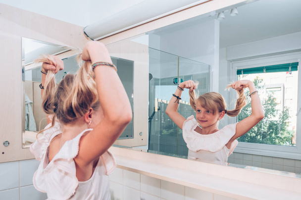linda criança loira no banheiro se divertindo com duas longas caudas loiras enquanto olha para o espelho
 - Foto, Imagem