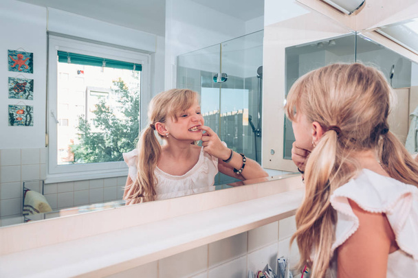 lovely blond child examining her healthy teeth in mirror in the bathroom  - Fotografie, Obrázek