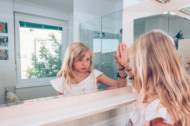 triest en serieus blond meisje kijken in de spiegel en het aanraken van het glas met de hand - Foto, afbeelding