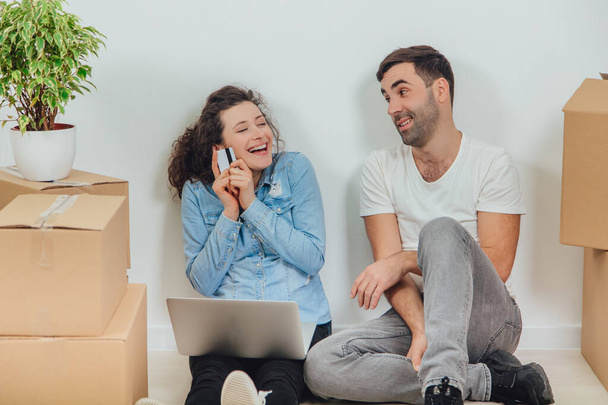 junges Paar mit Laptop in der Wohnung, auf dem Boden sitzend, im Internet surfend, auf der Suche nach neuen Möbeln. Frau ist überglücklich und hält Kreditkarte in der Hand. - Foto, Bild