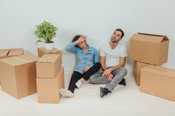 The process of moving into new house is totally exhausting. Lovely young couple wife and husband are sitting on the floor in new apartment among carton boxes. - Фото, зображення