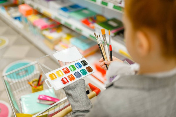 Little schoolgirl with a cart choosing watercolor paints, shopping in stationery store. Female child buying office supplies in shop, schoolchild in supermarket - Фото, изображение