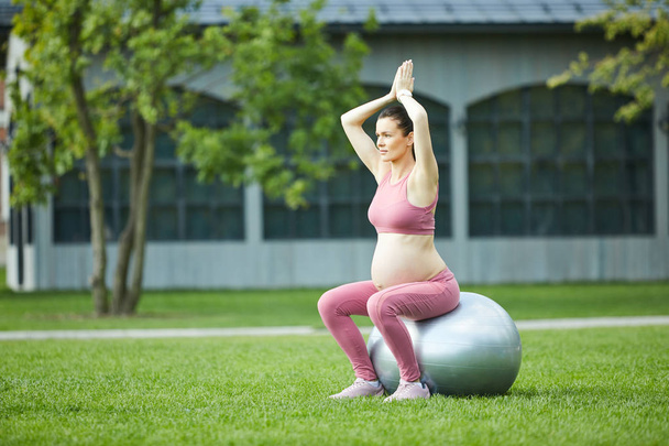 Jovem grávida em roupas esportivas rosa levantou os braços e meditando sobre a bola de fitness ao ar livre
 - Foto, Imagem