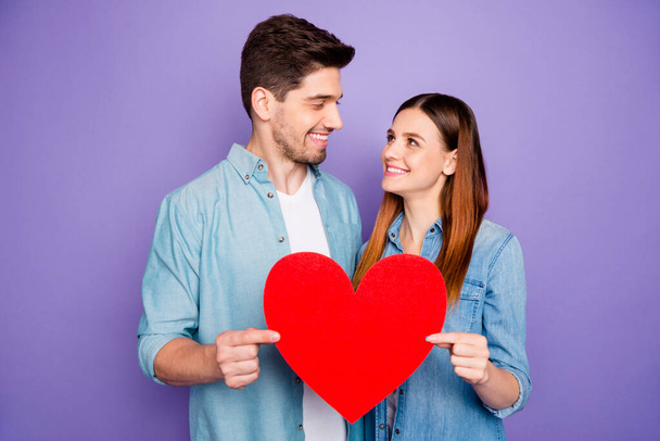 Portrait of romantic lovely spouses students hold paper card red big heart sign of their romance enjoy date 14-february holidays wear denim jeans shirt isolated over purple violet color background - Foto, imagen