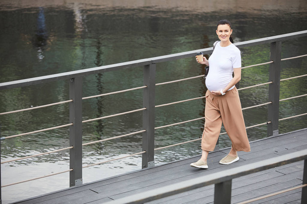 Πορτρέτο της νεαρής εγκύου που χαμογελά στην κάμερα πίνοντας καφέ και περπατώντας κατά μήκος της γέφυρας σε εξωτερικούς χώρους - Φωτογραφία, εικόνα