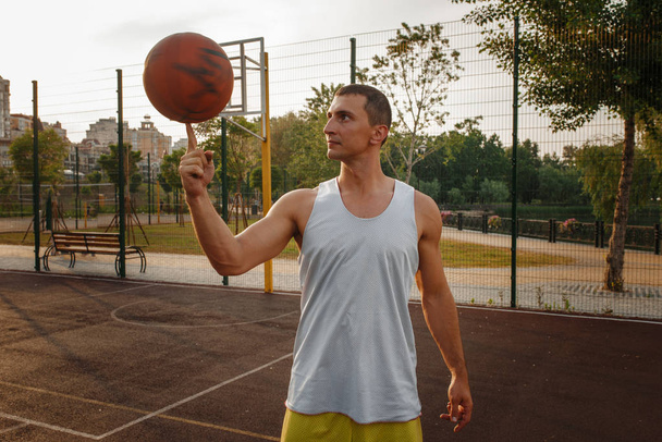 Мужчина-баскетболист крутит мяч на пальце на открытой площадке. Мужчина в спортивной одежде на тренировке по стритболу, летний стадион
 - Фото, изображение