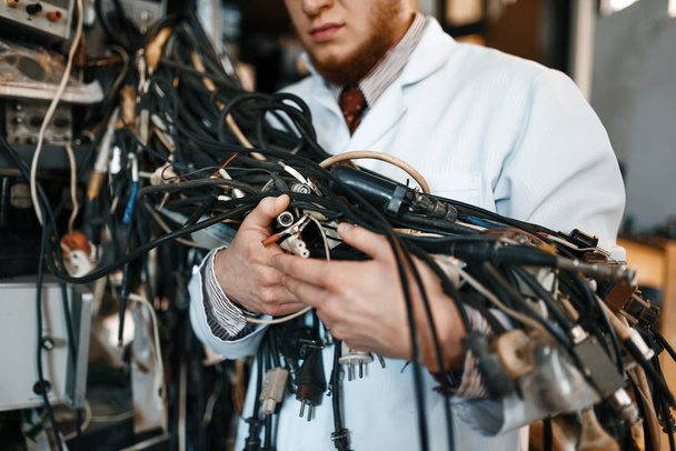 Ein seltsamer Ingenieur hält ein Bündel Drähte mit verschiedenen Steckern im Labor. Elektrische Prüfwerkzeuge im Hintergrund. Laborausstattung, technische Werkstatt - Foto, Bild