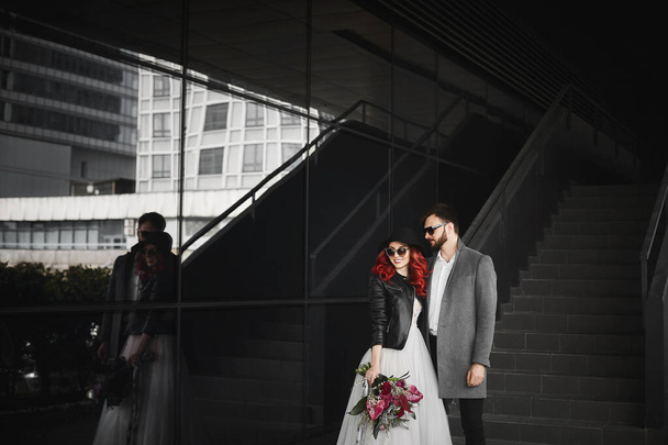 Beautiful couple of lovers posing outdoors during wedding photoshoot. Slim young woman with red hair in modish leather jacket and wedding dress and handsome bearded man in the coat. - Foto, Bild