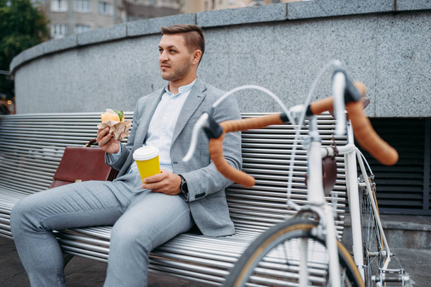 Businessman with cycle eats lunch on the bench at the office building in downtown. Business person riding on eco transport on city street, urban style - 写真・画像