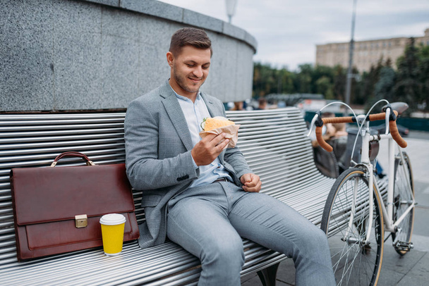 Un homme d'affaires à vélo mange le déjeuner sur le banc de l'immeuble de bureaux du centre-ville. Homme d'affaires à cheval sur l'éco transport sur la rue de la ville, style urbain
 - Photo, image