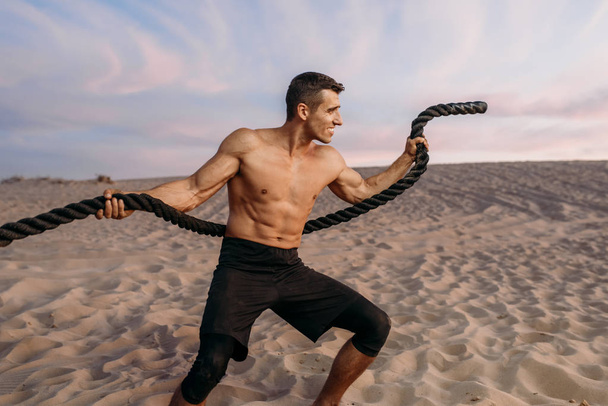 Мускулистый человек делает упражнения с веревкой в пустыне в солнечный день. Сильная мотивация в спорте, сильные тренировки на открытом воздухе
 - Фото, изображение