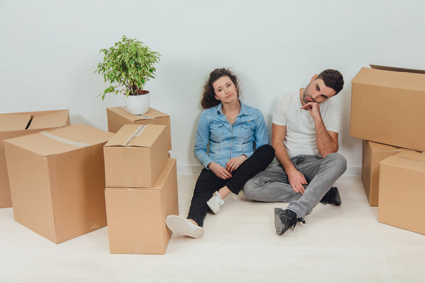 The process of moving into new house is totally exhausting. Lovely young couple wife and husband are sitting on the floor in new apartment among carton boxes. - Φωτογραφία, εικόνα