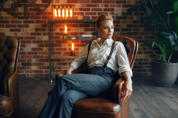Elegancka kobieta biznesu w ścisłej odzieży pozuje na skórzanej kanapie w studio, moda retro, styl gangsterski, mafia. - Zdjęcie, obraz