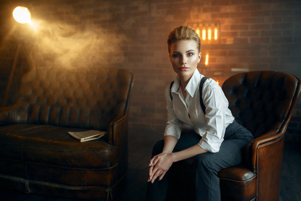 Elegant business woman in strict clothes poses on leather couch in studio, retro fashion, gangster style, mafia. Vintage lady in office with brick walls - 写真・画像