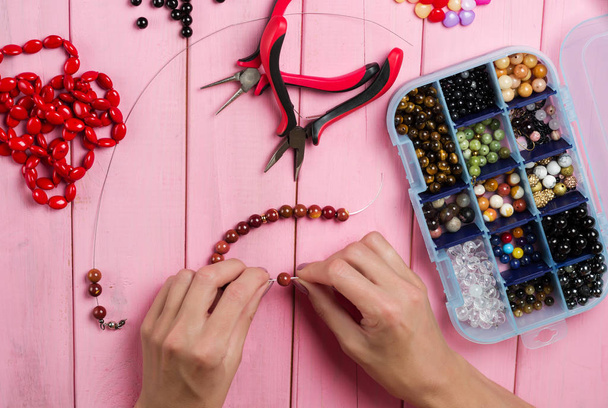 Jewelry making. Making bracelet of colorful beads. Female hands with a tool on a pink wooden table.  - Photo, Image