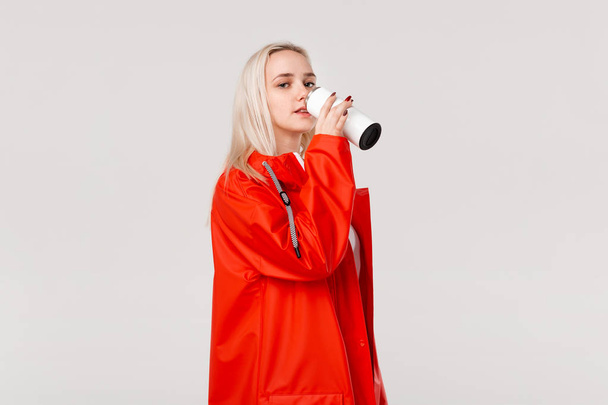 Blond žena v červeném plášti drží bílý termální hrnek s teplým nápojem izolované přes bílé pozadí. Připravte se na deštivé chladné dny - Fotografie, Obrázek