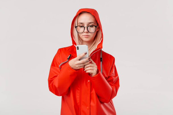 Blond woman in a red raincoat with a hood checking smartphone for notifications isolated over white background. Going for a walk on a rainy cold day - Fotó, kép
