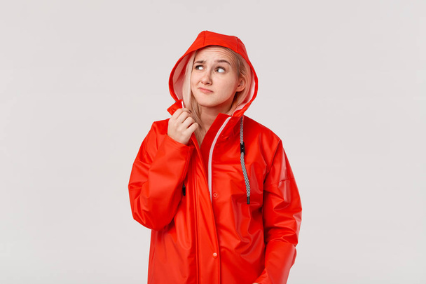 赤いレインコートを着たブロンドの女の子は、白い背景の上に孤立した雨が立ち始めたかどうかを確認するフードを持っています。寒い悪天候の準備はできていますか? - 写真・画像