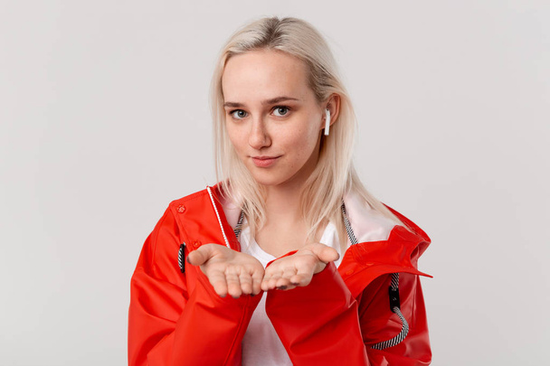 Smiling blond girl with wireless earbuds wearing red raincoat with hood holding palms of hands for avertisement isolated over white background. Place for advertising - Valokuva, kuva