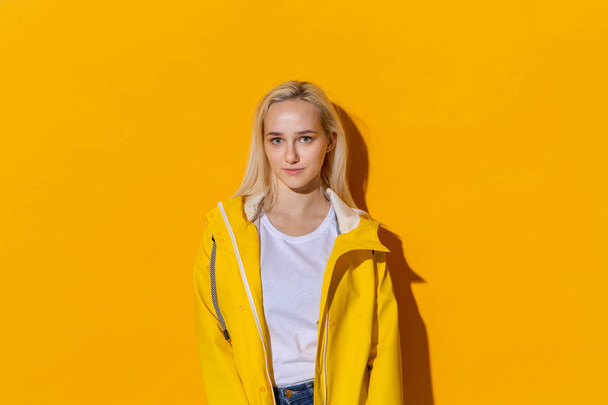 Chica rubia con un impermeable amarillo y una camiseta blanca de pie aislada sobre fondo amarillo. Ser brillante en el mal tiempo
. - Foto, imagen