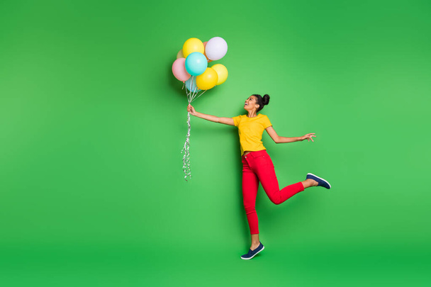 Full body profile photo of pretty millennial dark skin lady holding air balloons going to fly up in sky wear casual yellow t-shirt red pants isolated green background - Photo, Image