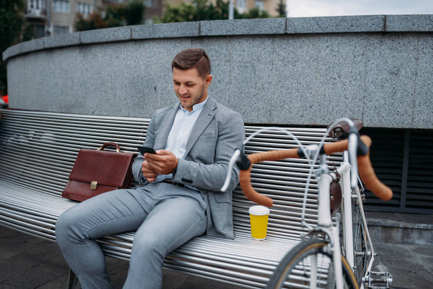 Businessman with bike talking by phone on the bench at the office building in downtown. Business person riding on eco transport on city street, urban style - Fotoğraf, Görsel