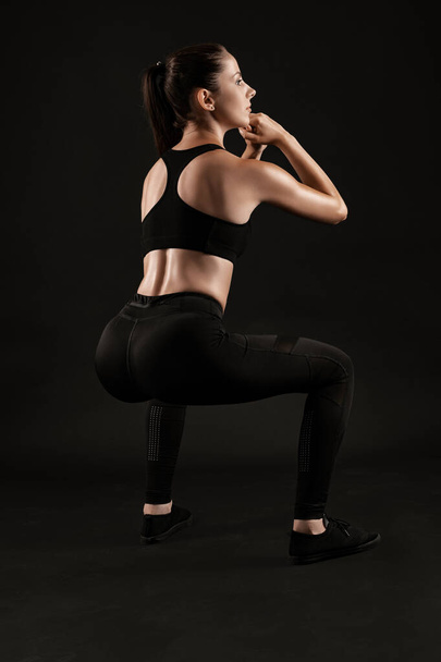 Brunette donna in leggings neri, top e sneakers è in posa su uno sfondo nero. Fitness, palestra, stile di vita sano. Lunghezza totale. - Foto, immagini