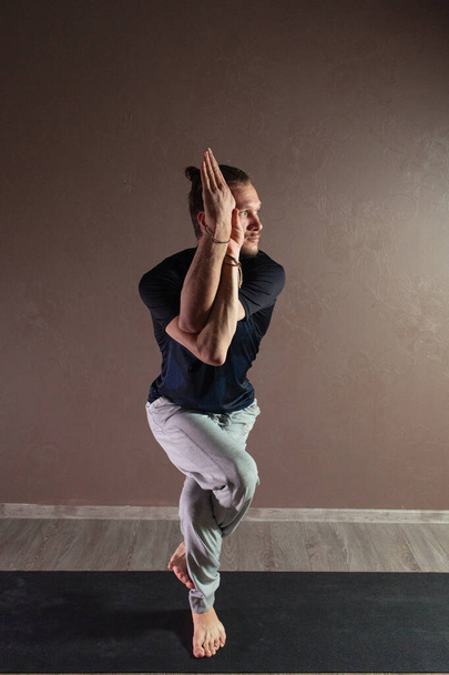 Young sporty man practicing yoga, meditating in Half Lotus pose, working out, wearing sportswear, indoor full length, gray yoga studio - Foto, afbeelding