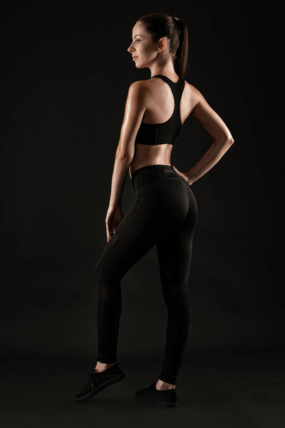 Mulher morena em leggings pretos, top e tênis está posando contra um fundo preto. Fitness, ginásio, conceito de estilo de vida saudável. Comprimento total. - Foto, Imagem