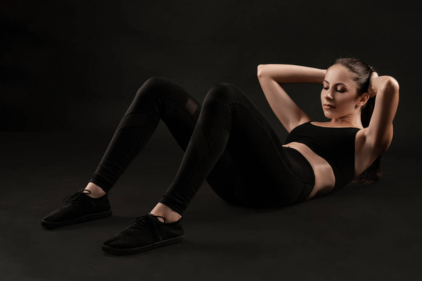 Brunette woman in black leggings, top and sneakers is posing against a black background. Fitness, gym, healthy lifestyle concept. Full length. - Zdjęcie, obraz