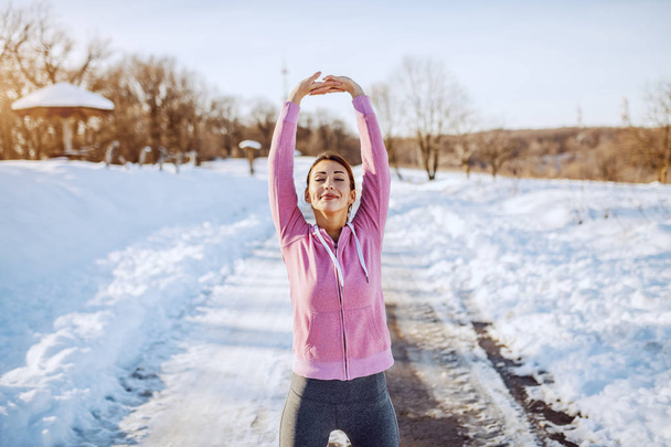 Three quarter length of beautiful Caucasian brunette in sportswear standing on country road and stretching arms before running. Wintertime. - 写真・画像