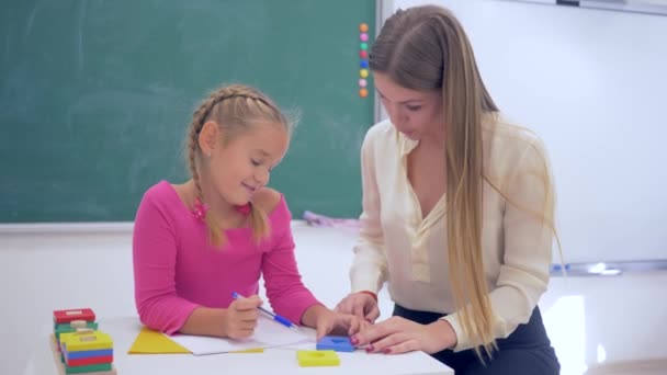 tutoring, female educator helps to scholar girl acquire information using plastic figures at table near blackboard in classroom of School - Footage, Video