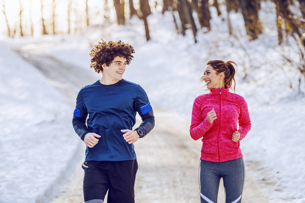 Happy smiling caucasian couple in sportswear running in nature while looking at each other. Winter time. - Photo, Image