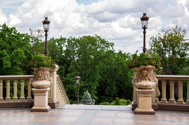pedestals with stone flowerpots with flowers at the stairs with railings and balustrades in the background stobes with lanterns green trees and cloudy sky. - Foto, Bild