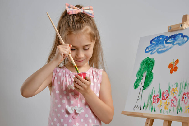 Pembe elbiseli küçük kız ressam sehpanın arkasında duruyor ve beyaz stüdyo arka planında izole edilmiş tuvalle resim yapıyor. Orta boy yakın çekim. - Fotoğraf, Görsel
