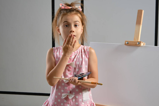 Μικρή καλλιτέχνης κορίτσι σε ένα ροζ φόρεμα στέκεται πίσω από καβαλέτο και ζωγραφική με πινέλο σε καμβά στο στούντιο τέχνης με λευκούς τοίχους. Μέτριο κοντινό πλάνο.. - Φωτογραφία, εικόνα