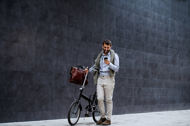 Sorrindo alegre homem de negócios da moda caucasiano usando telefone inteligente e empurrando sua bicicleta. No fundo é parede cinza
. - Foto, Imagem
