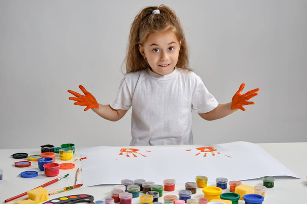 Κοριτσάκι με λευκό μπλουζάκι που κάθεται στο τραπέζι με τον whatman και πολύχρωμες μπογιές, δείχνοντας τα βαμμένα της χέρια. Απομονωμένο σε λευκό. Μέσο κοντινό πλάνο. - Φωτογραφία, εικόνα
