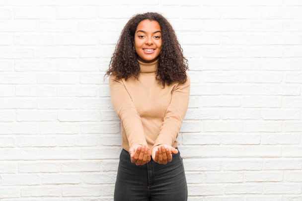 young black woman smiling happily with friendly, confident, positive look, offering and showing an object or concept against brick wall - Foto, Bild