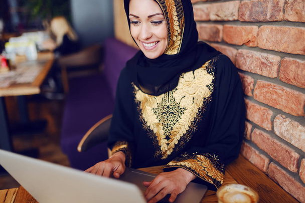 Impressionante mulher de negócios muçulmana vestida com o desgaste tradicional sentado no café e usando laptop. Na mesa estão laptop e café
. - Foto, Imagem