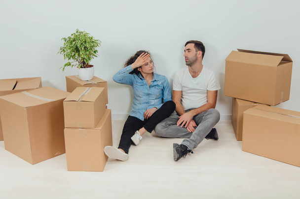 The process of moving into new house is totally exhausting. Lovely young couple wife and husband are sitting on the floor in new apartment among carton boxes. - Fotografie, Obrázek