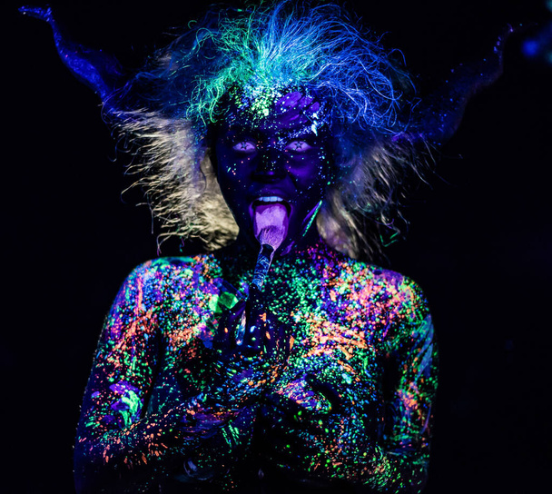 The girl in the image of the devil, contact lenses and horns. Woman paints her tongue in ultraviolet paint. fluorescent powder. Body art glowing in ultraviolet light. Stars in the eyes. - Fotó, kép