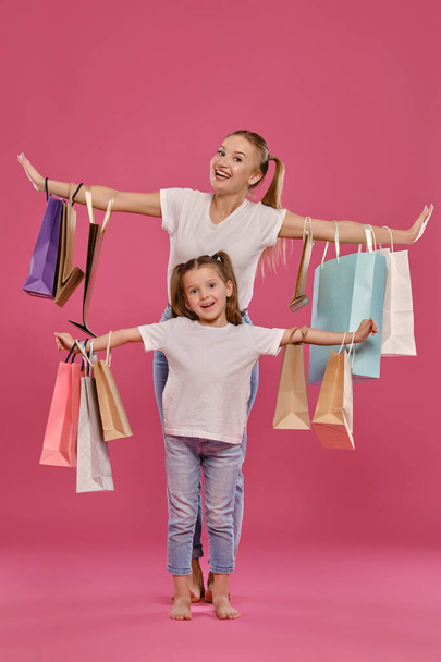 Mutter und Tochter mit Pferdeschwanz, gekleidet in weiße T-Shirts und blaue Jeans posieren vor rosa Hintergrund mit Paketen in der Hand. Volle Länge. - Foto, Bild